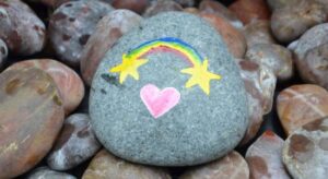 Bunte bemalte Steine mit einem Regenbogen, Sternen und einem Herz auf einem Haufen grauer Kieselsteine.