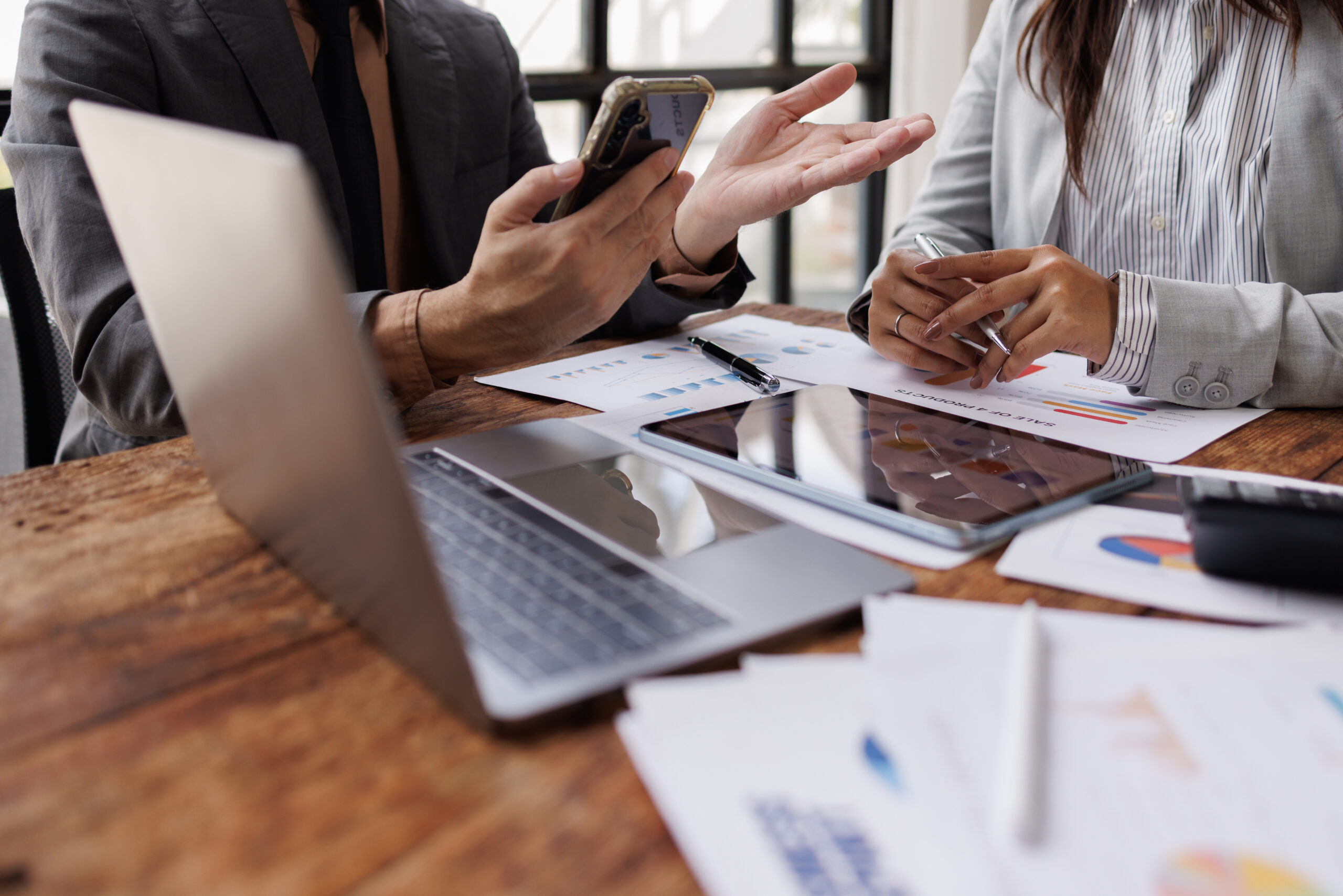 Zwei Personen in Businesskleidung diskutieren &uuml;ber Dokumente und nutzen ein Smartphone und ein Laptop auf einem Holztisch.