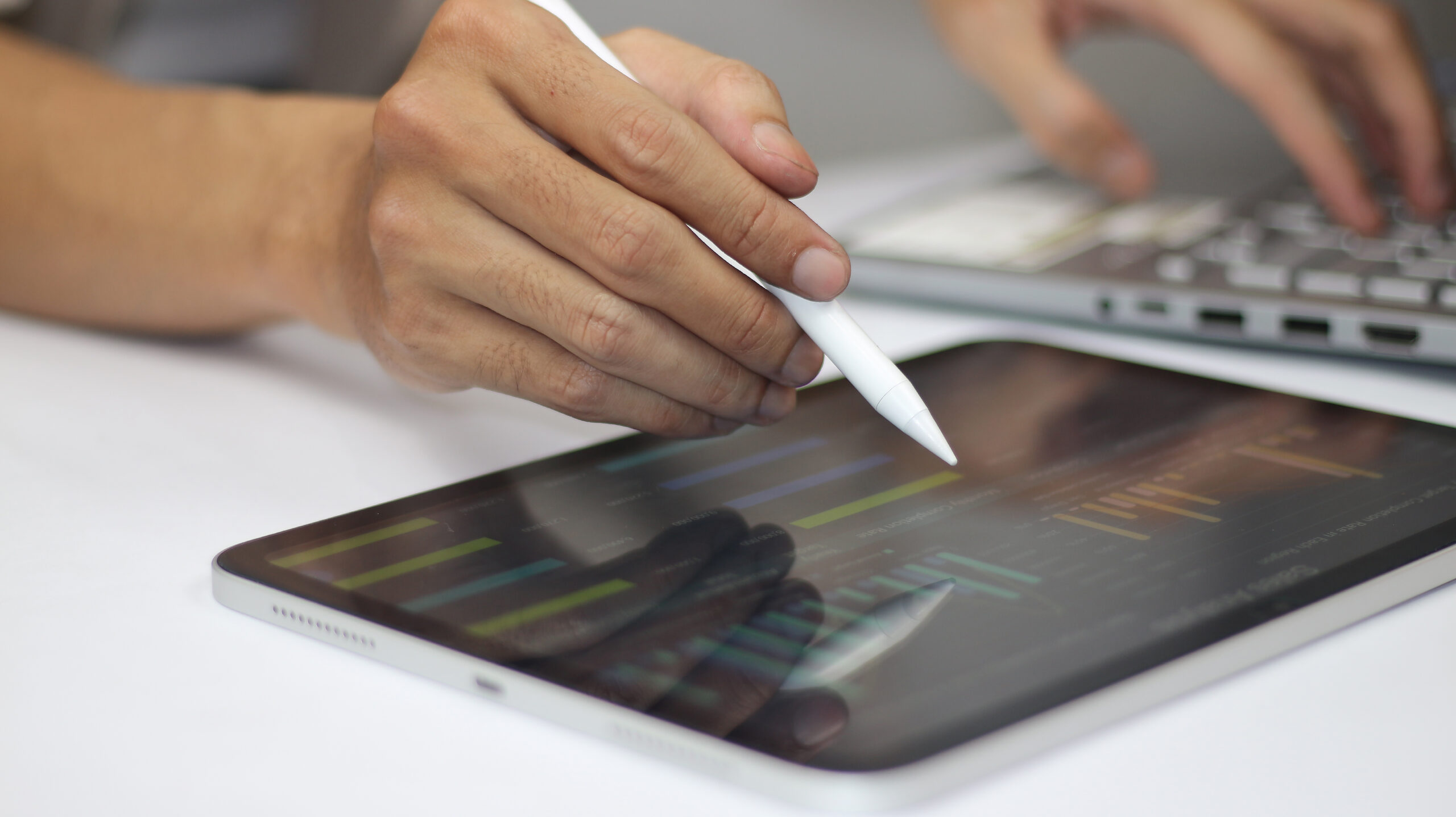 Eine Hand mit einem Stylus verwendet ein Tablet, w&auml;hrend ein Laptop im Hintergrund sichtbar ist.