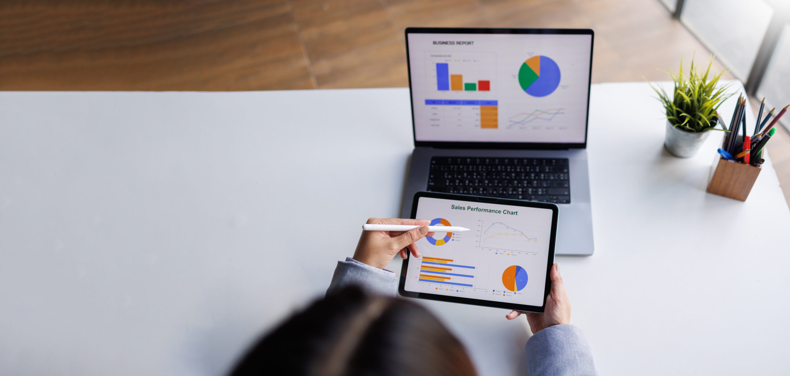 Eine Person analysiert Verkaufsdaten auf einem Tablet, w&auml;hrend auf einem Laptop Diagramme und Grafiken angezeigt werden.
